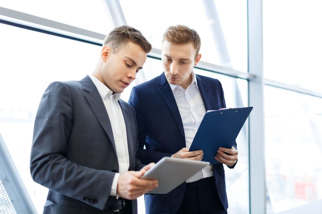 Dos hombres de negocios consultando documentos y una tableta digital en un entorno de oficina moderno con grandes ventanas.