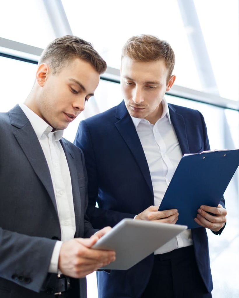 Dos hombres de negocios consultando documentos y una tableta digital en un entorno de oficina moderno con grandes ventanas.